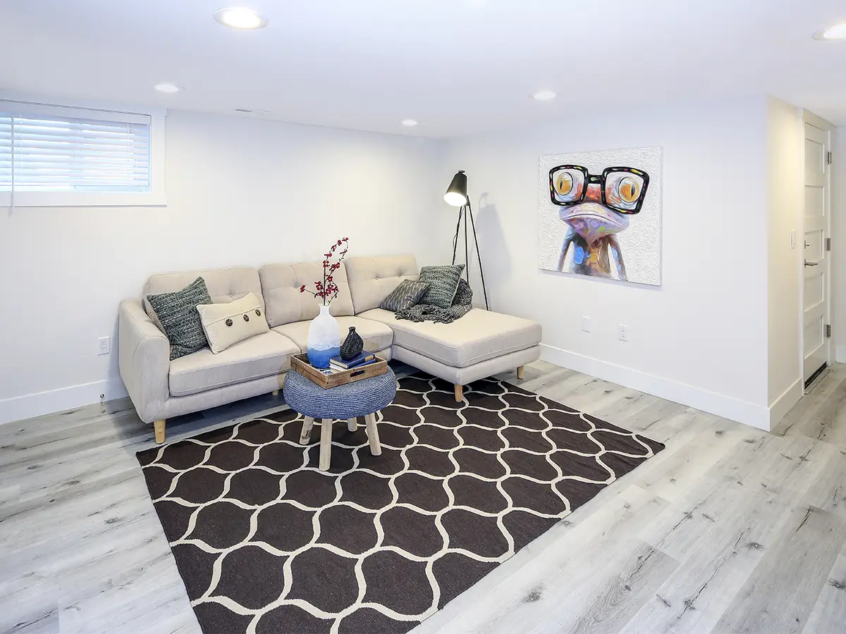 Basement featuring a stylish white-gray sofa, juxtaposed against a gray wooden floor and a patterned carpet. The walls are adorned in elegant white tones, accentuated by spotlights that illuminate the space. This combination of contemporary elements and subtle patterns creates a sophisticated and inviting ambiance, perfect for relaxation or entertaining guests in style