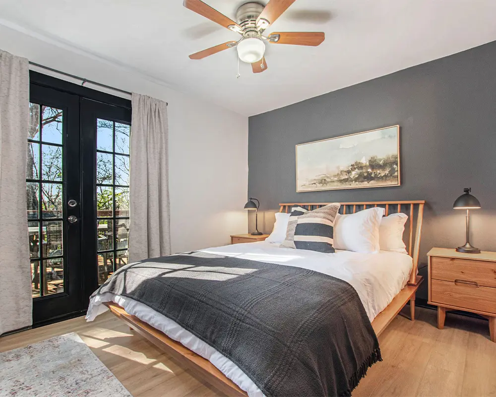 Tranquil bedroom featuring a wooden bed and flooring, complemented by one accent wall in dark gray and the remaining walls in crisp white. The striking contrast adds depth and character to the space. A black door leads to an outdoor balcony, offering a refreshing escape and a glimpse of the outside world. The room's harmonious blend of natural elements and contemporary design invites serenity and relaxation, making it a cozy sanctuary within the home