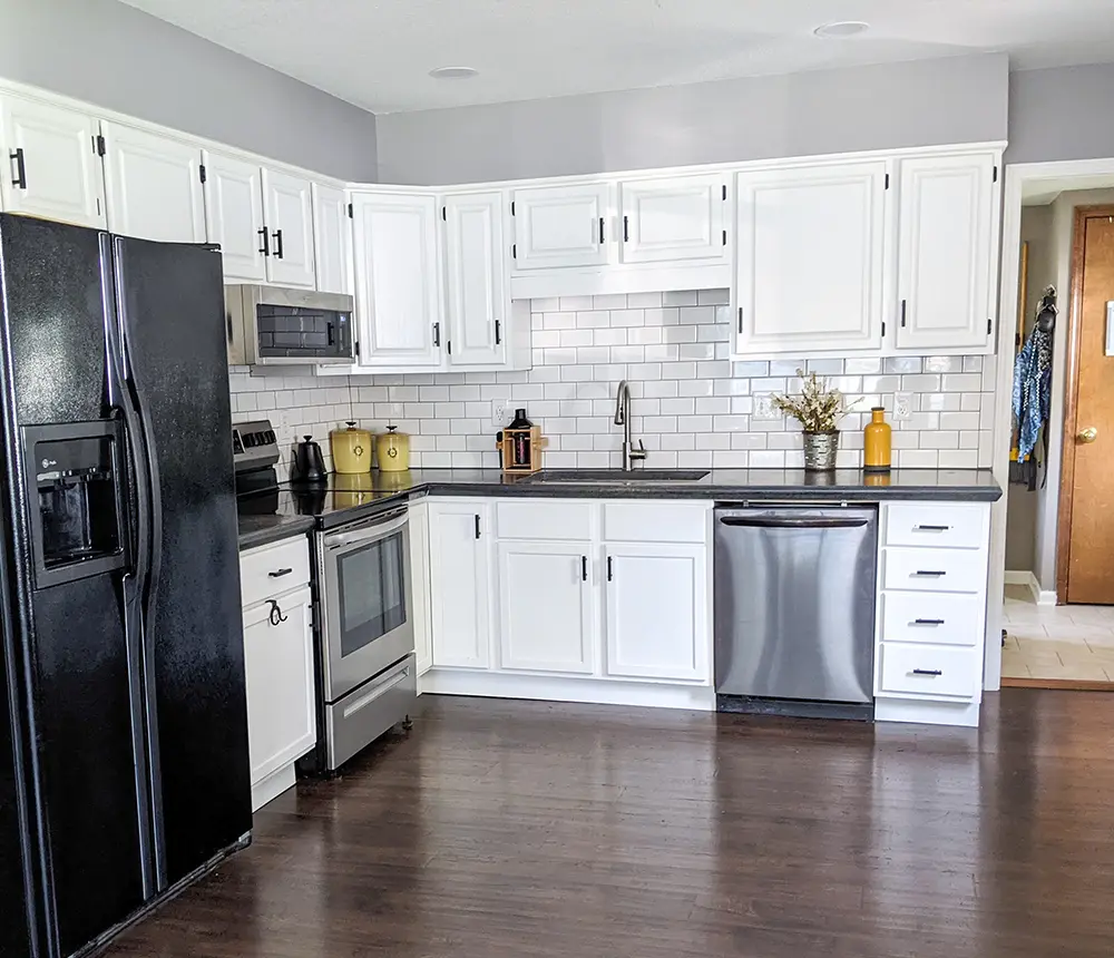 Newly remodeled kitchen boasting dark wooden flooring and pristine white cabinets, providing a striking contrast. Enhanced with sleek black fridge and countertops, infusing a modern essence into the space. The fusion of dark and light elements creates a sophisticated aesthetic, marrying elegance with functionality in this contemporary kitchen design.