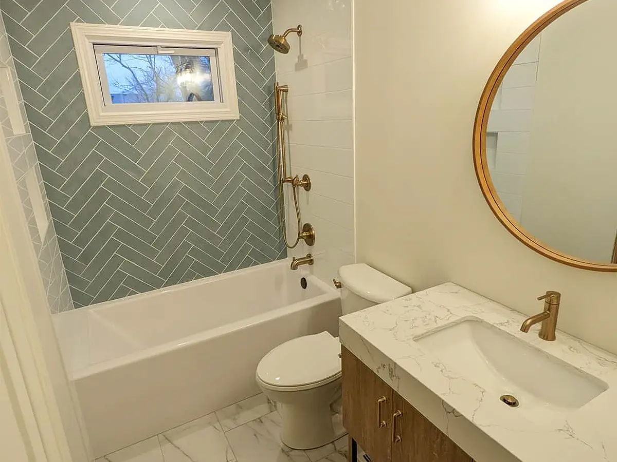 Compact bathroom featuring white tones with a modern touch. Gray patterned tiles adorn the walls, accentuating the contemporary design. A wooden cabinet with a sink, complemented by golden fixtures, adds a luxurious feel. Stylish and functional space for daily routines