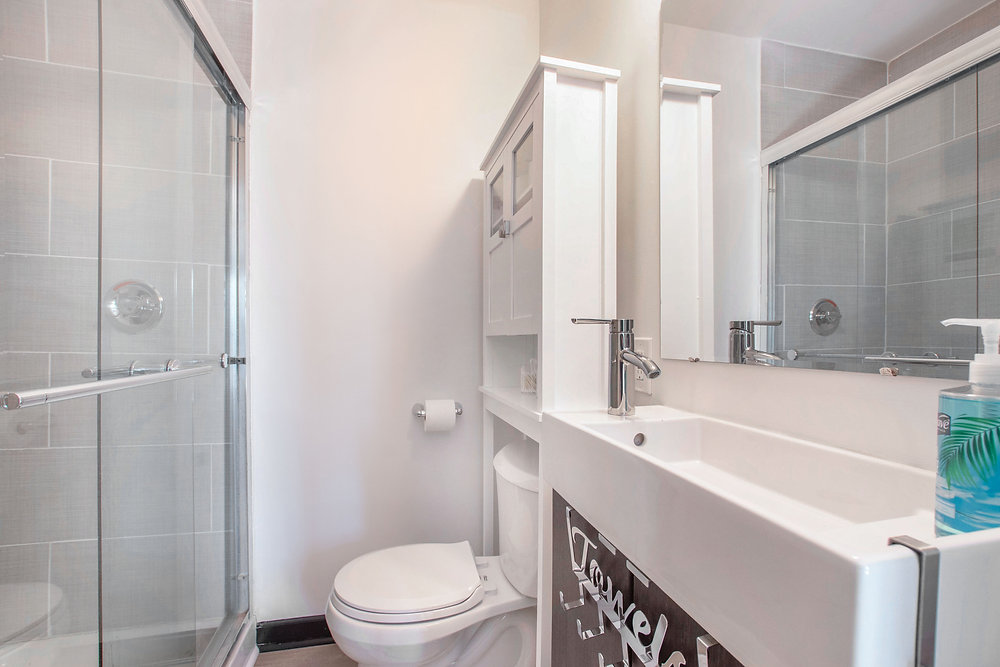 A small white-themed bathroom exuding cleanliness and simplicity. The space features a compact layout with white tiles, a pedestal sink, and minimalist fixtures, creating an airy and refreshing atmosphere. The cohesive white color scheme enhances the sense of cleanliness and brightness, making the bathroom an inviting and refreshing space for daily use.