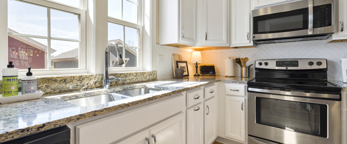 Bright kitchen with white cabinets, granite countertops, stainless steel appliances, and a double sink under large windows.
