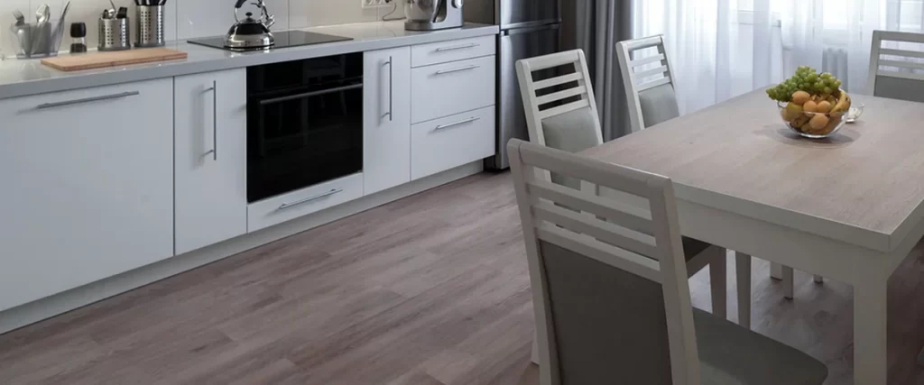 Modern kitchen with vinyl flooring, sleek white cabinetry, stainless steel appliances, and a dining table near large windows.