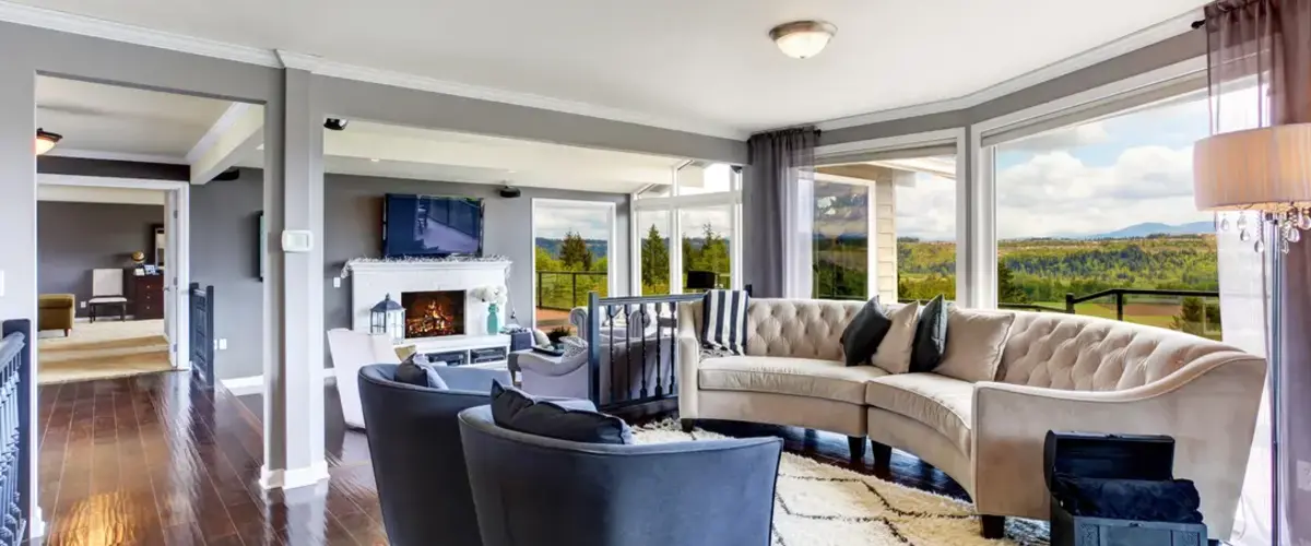 A bright, luxurious living room featuring a curved beige sofa and floor-to-ceiling windows that offer a panoramic view of the landscape and natural light.