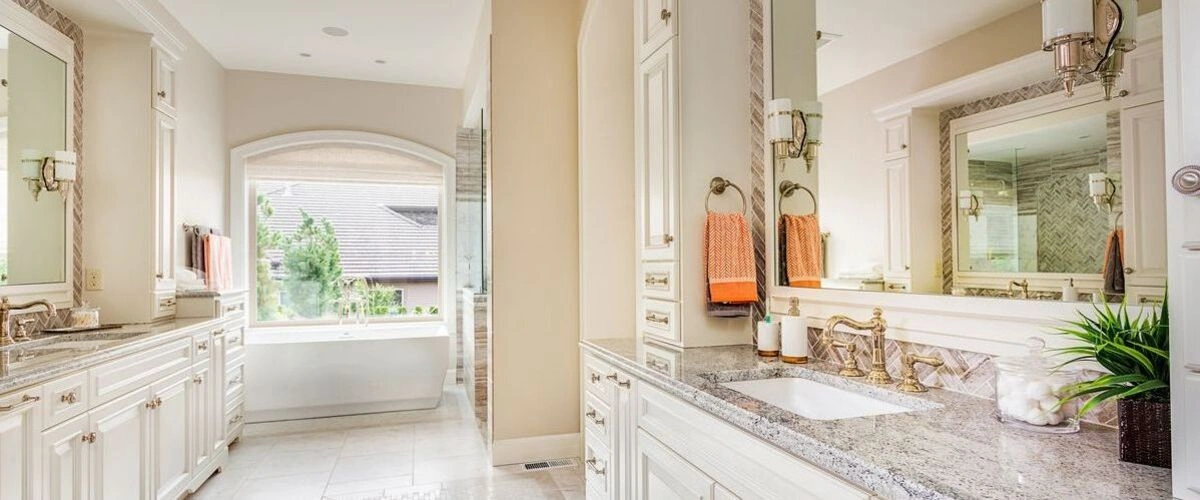 Bright and elegant washroom interior showcasing a long double vanity with white cabinetry, beige granite surfaces, and a soaking tub under a window.