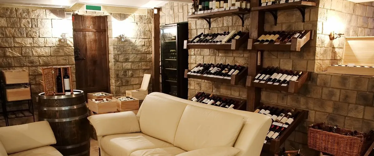 A rustic luxury basement wine cellar with natural stone walls, custom wooden wine racks filled with bottles, a white leather sofa, and decorative wine barrels.