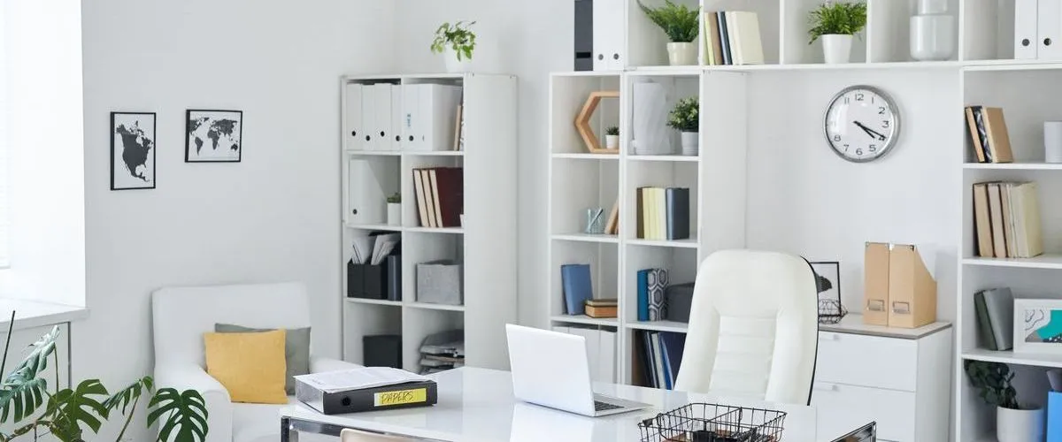 A bright and airy modern home office located in a finished basement featuring white shelving units, a clean white desk with a laptop, and a comfortable ergonomic chair.