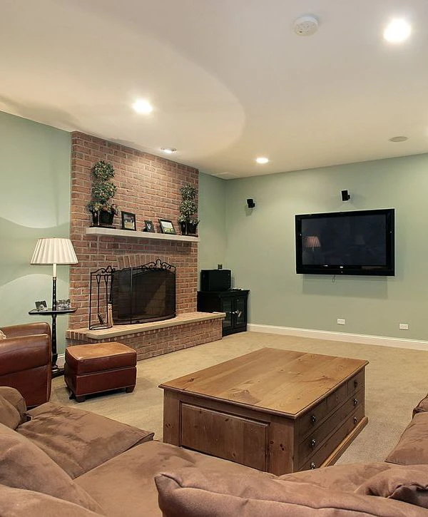 A cozy finished basement living area featuring a custom brick fireplace, soft green walls, a flat-screen TV, and brown seating, designed by top basement remodeling companies in Raymore, MO.