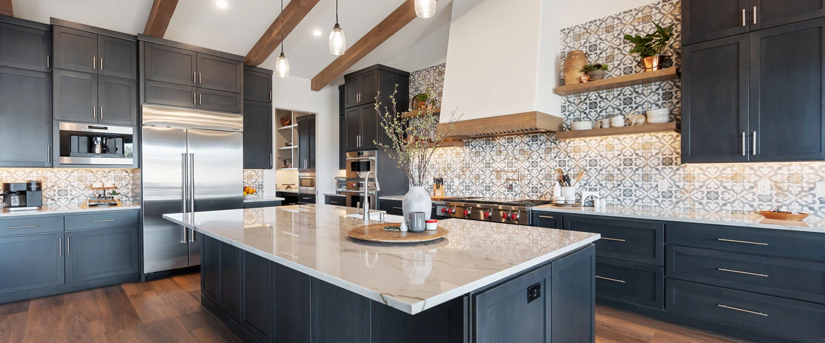A spacious modern kitchen remodel featuring dark charcoal cabinetry, a large white marble island, patterned tile backsplash, and rustic wood ceiling beams.