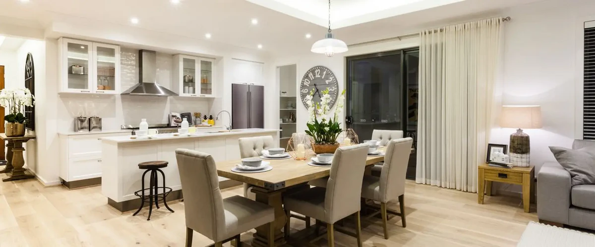 An open-concept living space featuring a wooden dining table with grey upholstered chairs leading into a bright white kitchen with a large island and stainless steel appliances.