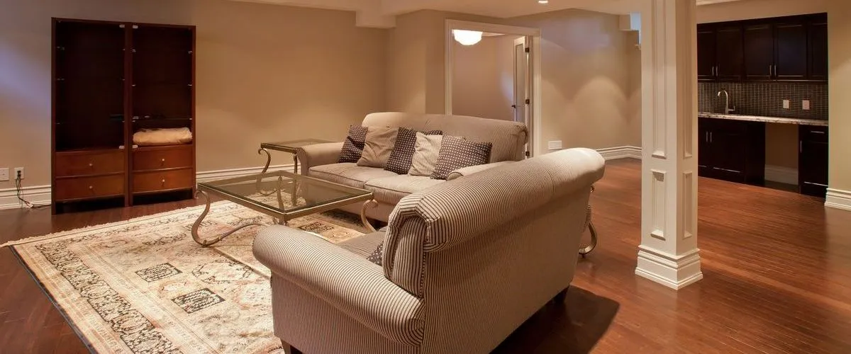 A spacious open-concept finished basement showcasing dark hardwood flooring, beige striped sofas, a glass coffee table, and a custom dark wood wet bar in the background.