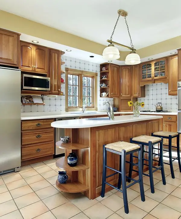 Traditional kitchen with warm wood cabinets, a white countertop island featuring rounded open shelves, and three dark blue woven stools, completed by one of the top kitchen remodeling companies in Raymore, MO.